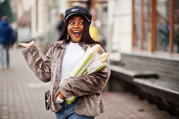 Modieuze Afro-Amerikaanse vrouw in zwarte pet en bont met twee stokbrood rollen bij de hand