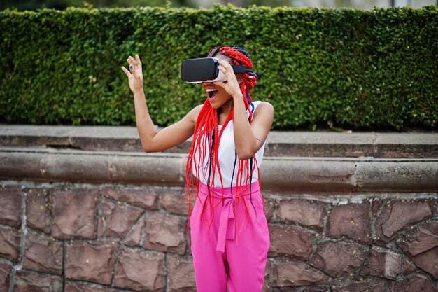 Modieuze Afro-Amerikaanse zakenvrouw in roze broek en rode dreadlocks met virtual reality-bril buiten