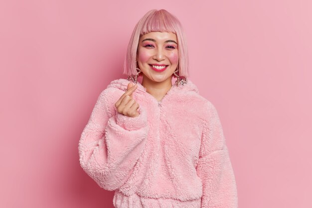 Modieuze Aziatische vrouw met lichte make-up maakt mini hart gebaar Koreaans als teken glimlach aangenaam heeft roze haar en bontjas vormt indoor jurken voor disco party. Lichaamstaal concept.