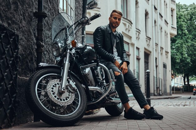 Modieuze biker gekleed in een zwart leren jack en jeans zittend op zijn retro motorfiets op oude Europa straat.