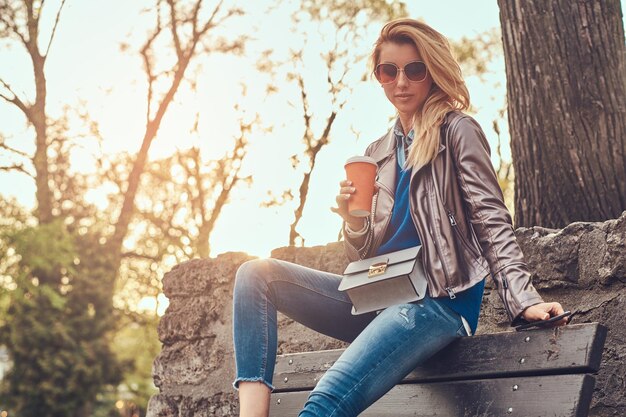 Modieuze blonde vrouw ontspant buiten en drinkt afhaalkoffie terwijl ze op de bank in het stadspark zit.
