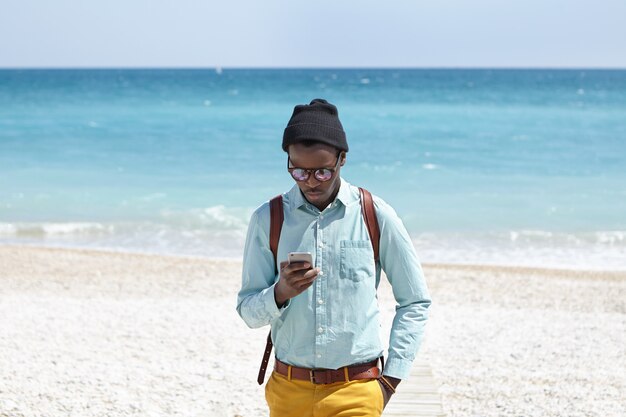 Modieuze jonge Afro-Amerikaanse mannelijke toerist met behulp van mobiele telefoon op woestijnstrand, het plaatsen van foto's van prachtig zeegezicht om hem heen via sociale media met azuurblauwe oceaan en blauwe lucht in horizon