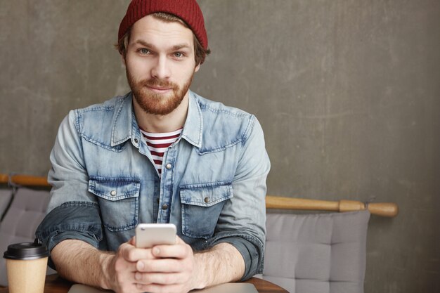 Modieuze jonge bebaarde man in hoed en spijkerblouse zittend aan cafétafel met papieren kopje verse koffie, met mobiele telefoon terwijl hij online een bericht verstuurt en op internet surft, met gratis wifi