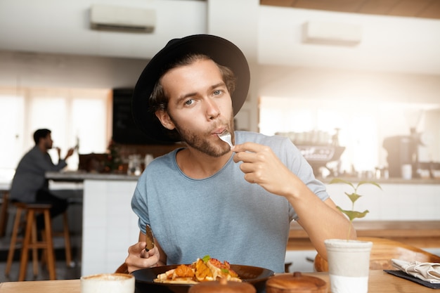 Modieuze jonge man genieten van lekker eten voor lunch zittend aan houten tafel van gezellig restaurant. Hongerige hipster die trendy zwarte hoed dragen die zijn honger stillen terwijl het hebben van maaltijd bij alleen cafetaria