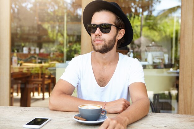 Modieuze jonge man in trendy zonnebril en wit V-hals shirt met rust op de stoep cafetaria, cappuccino drinken