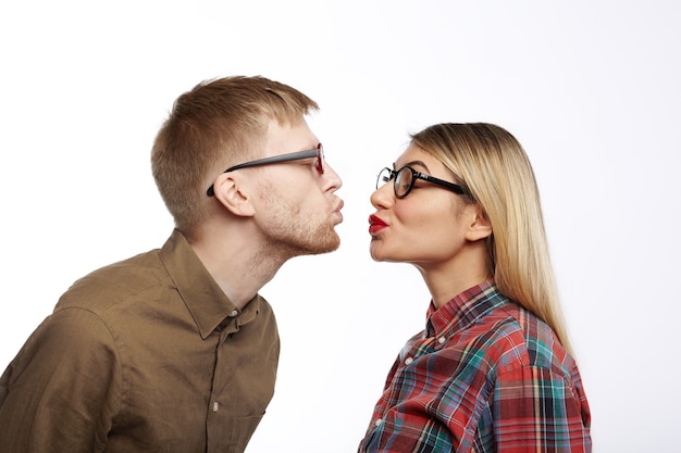 Modieuze jonge man met stoppels en mooie stijlvolle vrouw pruilen hun lippen en ogen sluiten, op het punt om te kussen. Zijwaarts portret van schattige zoete hipster paar verliefd zoenen voorbereiden