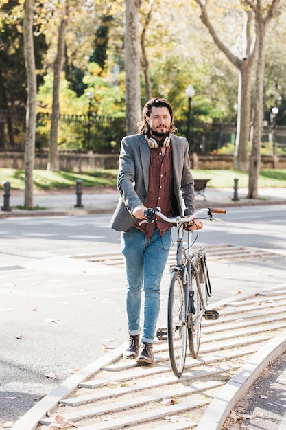 Gratis foto modieuze jonge mens met hoofdtelefoon rond zijn hals die met zijn fiets op stoep lopen