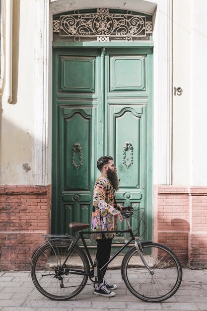 Modieuze jonge mens met zijn fiets die zich voor groene deur bevindt