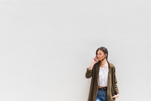 Modieuze jonge vrouw die zich tegen witte muur bevindt die op mobiele telefoon spreekt