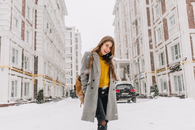 Modieuze jonge vrouw in jas met rugzak lopen op straat in de grote stad in sneeuwt tijd. vrolijke stemming, sneeuwval, wachten op kerstmis, positiviteit uiten, ware emoties.