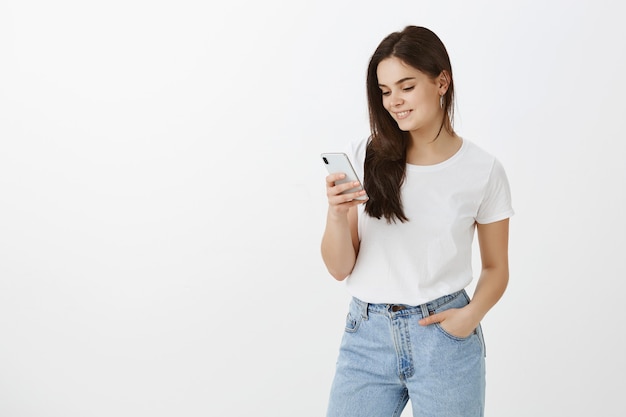 Modieuze jonge vrouw poseren met haar telefoon tegen witte muur