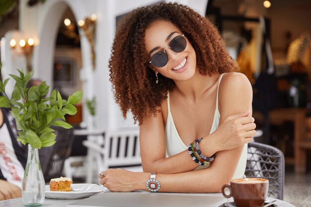 Modieuze lachende blij Afro-Amerikaanse vrouw in trendy zonnebril, drinkt koffie of latte, eet zoete cake, geniet van vrije tijd in café