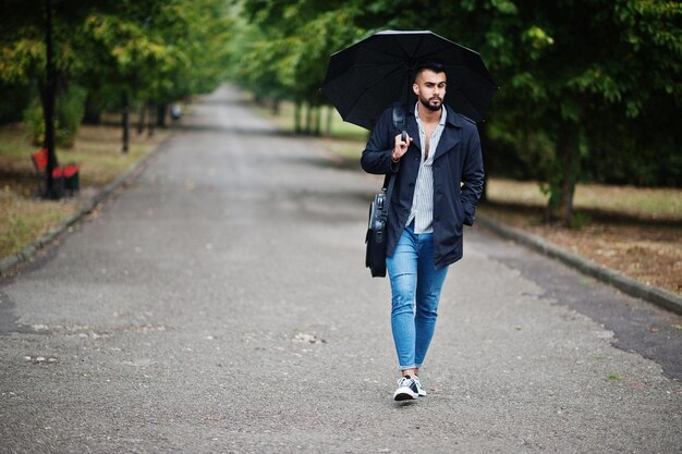 Modieuze lange Arabische baard man draagt zwarte jas met paraplu en tas geposeerd op regenweer dag