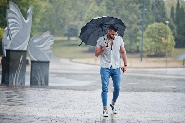 Modieuze lange Arabische baardman draagt een shirtjeans en zonnebril met paraplu bij regen op het parkplein