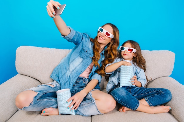 Modieuze mooie moeder in jeans kleding selfie portret met haar jonge dochter op bank geïsoleerd op blauwe achtergrond. 3d-bril dragen, popcorn eten, samen film kijken