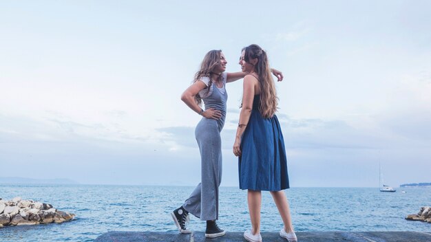 Modieuze twee jonge vrouwelijke vrienden die zich tegen blauwe overzees en hemel bevinden