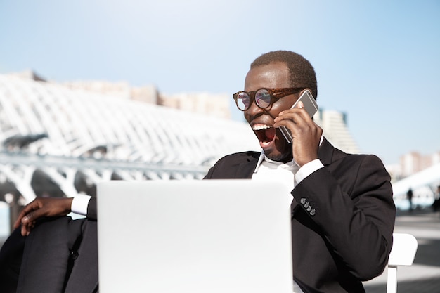 Moe geeuwende jonge afro-amerikaanse bankier gekleed formeel zittend aan terras tafel voor notebookcomputer en praten op mobiele telefoon terwijl hij wacht op zijn lunch, verveeld slaperig blik