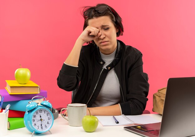 Moe jong student meisje bril op hoofd zit aan bureau met universitaire tools huiswerk wrijven haar oog geïsoleerd op roze achtergrond