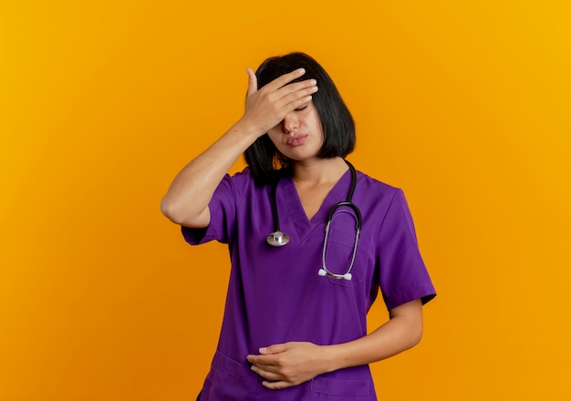 Moe jonge brunette vrouwelijke arts in uniform met stethoscoop legt hand op voorhoofd