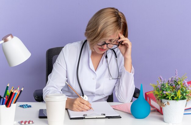 Moe jonge vrouwelijke arts dragen medische gewaad met stethoscoop en bril zit aan tafel met medische hulpmiddelen schrijven iets op Klembord geïsoleerd op blauwe achtergrond