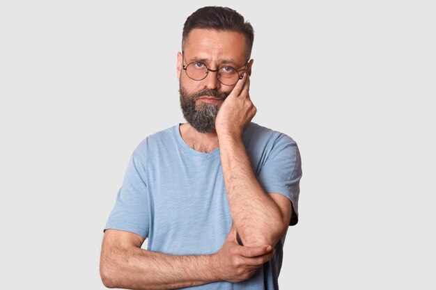 moe kalm man staande geïsoleerd op wit, zijn gezicht aan te raken met een hand, diep van streek, het dragen van t-shirt en trendy bril.