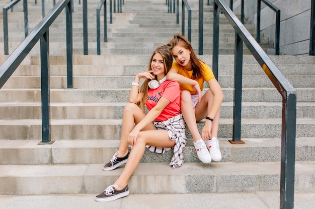 Moe maar tevreden brunette meisje in trendy witte sneakers ontspannen op stenen trappen met beste vriend