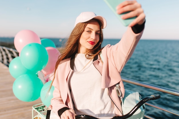 Moe meisje met een stijlvolle make-up wordt gefotografeerd op de achtergrond van de zee na een fietstocht in de ochtend langs de pier.