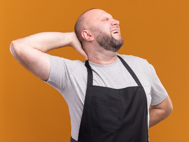 Moe met gesloten ogen Slavische mannelijke kapper van middelbare leeftijd in uniform hand zetten achter hoofd geïsoleerd op oranje muur