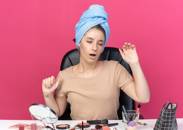 Moe met gesloten ogen zit jong mooi meisje aan tafel met make-up tools gewikkeld haar in handdoek geïsoleerd op roze achtergrond