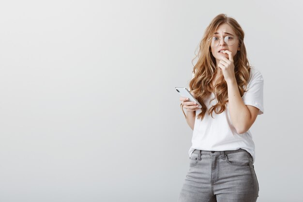 Moeder belt om te kijken of dochter thuis is. Portret van angstige knappe blanke vrouw in trendy bril, bijtende vinger en fronst terwijl ze zenuwachtig is, smartphone houdt over grijze muur