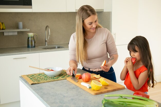 Moeder die dochter geeft om appelschijf te proeven tijdens het koken van salade. Meisje en haar moeder samen koken, vers fruit en groenten snijden op snijplank in de keuken. Familie koken concept