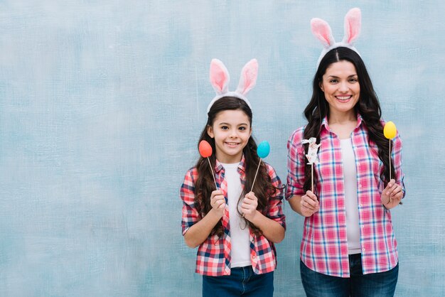 Moeder en dochter die konijntjesoren dragen die paaseieren en konijntjessteunen houden