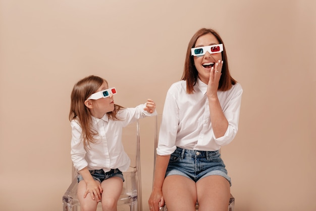 Moeder en dochter op zoek naar elkaar en lachend in 3d-bril naar camera op bank geïsoleerd op beige achtergrond.