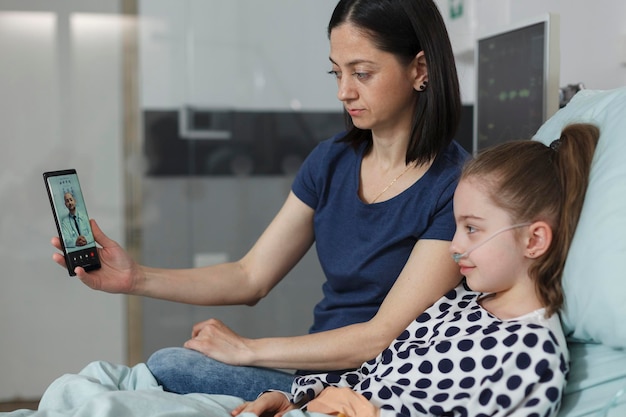 Moeder en zieke dochter praten met arts over telehealth telefoon videogesprek terwijl ze op de kinderafdeling van het ziekenhuis zitten. Ziek kind en ouder bespreken met arts op smartphone virtueel online gesprek