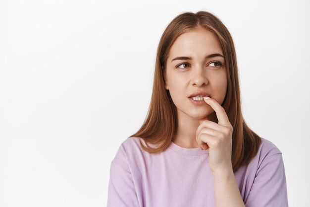 Moet nadenken. Jonge vrouw bijt in haar vinger terwijl ze nadenkt, nadenkend wegkijkt, berekenend in gedachten, besluit, staande tegen een witte achtergrond