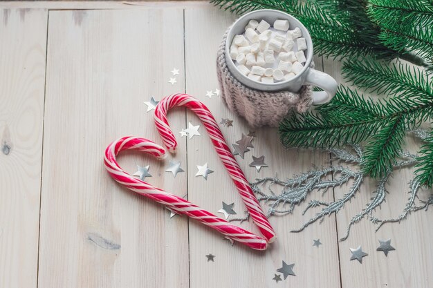 Mok van marshmallows en zuurstokken omgeven door kerstversiering op een houten tafel