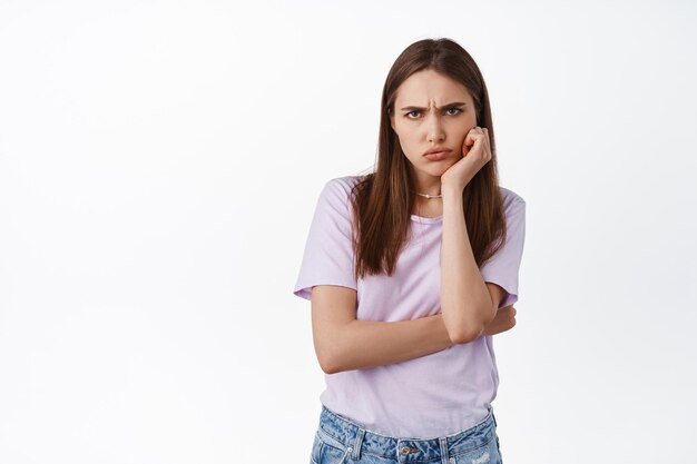 Mokkende boze vriendin fronsen, pruilen en beledigd kijken, beledigd zijn, overstuur zijn over iets, ontevreden grimassen, staande tegen een witte achtergrond