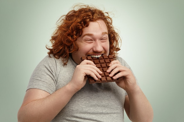 Mollige grappige roodharige jonge blanke man met krullend haar bijt groot van chocolade