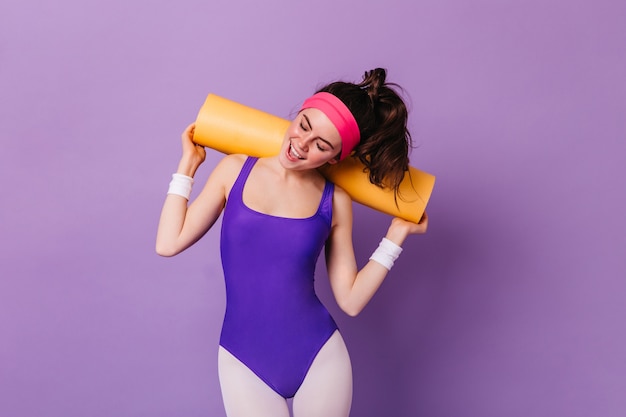 Momentopname van aantrekkelijke vrouw in sportkleding in stijl van de jaren 80, poseren met aerobicsmat op paarse muur