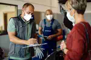 Gratis foto monteur met gezichtsmasker die rapporten doorneemt terwijl hij met de klant in de autoreparatiewerkplaats praat