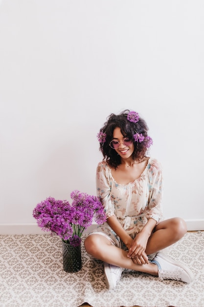 Mooi afrikaans meisje in schattige zomerjurk zit in de buurt van vaas met alliums. indoor portret van brunette zwarte dame thuis koelen.