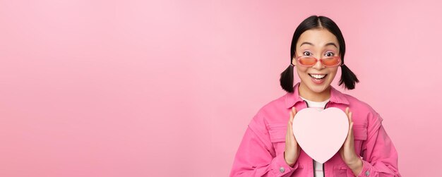 Mooi aziatisch meisje lacht blij met het tonen van een hartgeschenkdoos en kijkt opgewonden naar de camera die over een roze romantische achtergrond staat