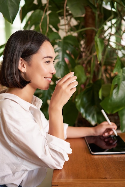 Mooi aziatisch meisje zit in café met kopje koffie tekenen op digitale tablet met grafische pen d