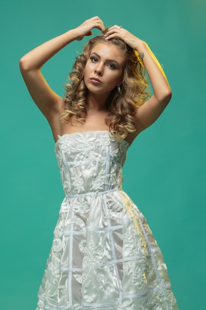 Gratis foto mooi blond meisje in een witte jurk die haar haar op een blauwe achtergrond verzamelt. foto van hoge kwaliteit