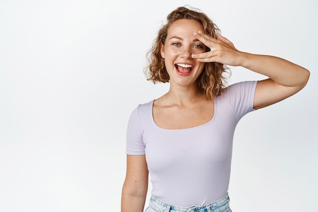 Mooi blond meisje met oog vrede gebaar, lachen en zorgeloos glimlachen, staande in tshirt op wit. ruimte kopiëren