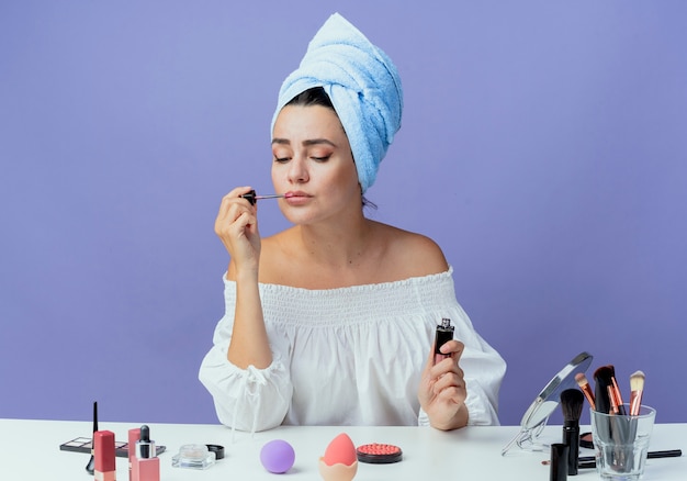 Mooi boos meisje gewikkeld haar handdoek zit aan tafel met make-up tools vasthouden en lipgloss toe te passen neerkijkt geïsoleerd op paarse muur