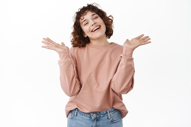 Mooi en gelukkig meisje spreidde handen zijwaarts, verrassingsgebaar, schouderophalend met onzorgvuldig en onbezorgd gezicht, staande in roze sweatshirt tegen witte achtergrond.