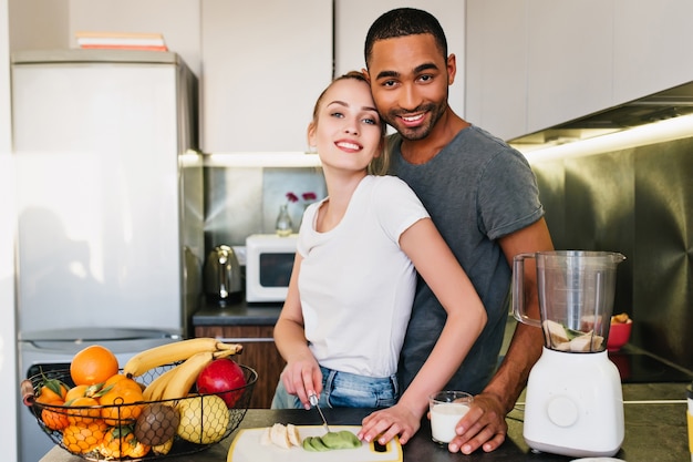 Mooi en paar kijken en glimlachen. Man en vrouw koken samen in de keuken. Blonde snijdt fruit. Liefhebbers in T-shirts met blije gezichten brengen thuis tijd samen door.