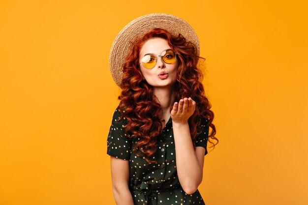 Mooi gembermeisje die luchtkus op gele achtergrond verzenden. Studio shot van krullende jonge vrouw in zonnebril en strooien hoed.