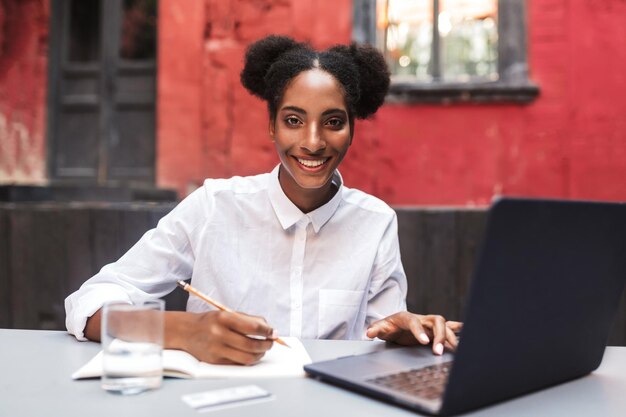 Mooi glimlachend Afrikaans meisje in een wit overhemd dat gelukkig in de camera kijkt terwijl ze met een laptop werkt op de gezellige binnenplaats van het café